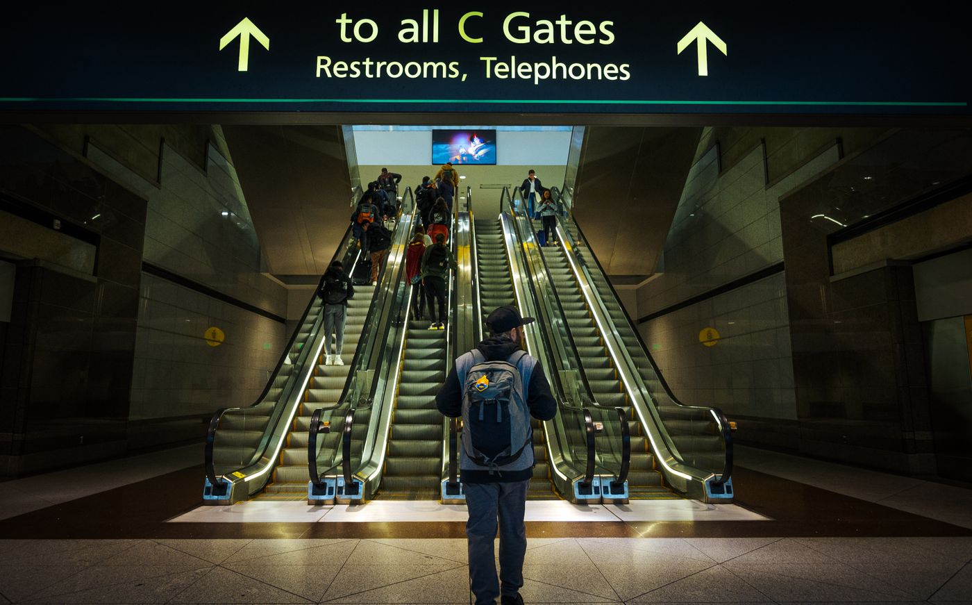 A traveller with a backpack walks towards escalators under a sign reading 'to all C Gates'